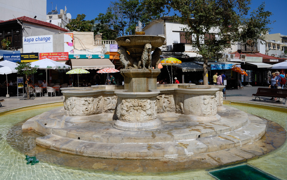 Heraklion, Der Morosinibrunnen (erbaut 1628) auf der von zahlreichen Straßencafés umgebenen Platia Venizelou bildet das Herz der Stadt. Der aus acht reliefgeschmückten Wasserbecken mit einer von vier steinernen Löwen getragenen Wasserschale in der Mitte bestehende Brunnen wurde über ein 15 km langes Aquädukt aus den Quellen von Archanes gespeist.